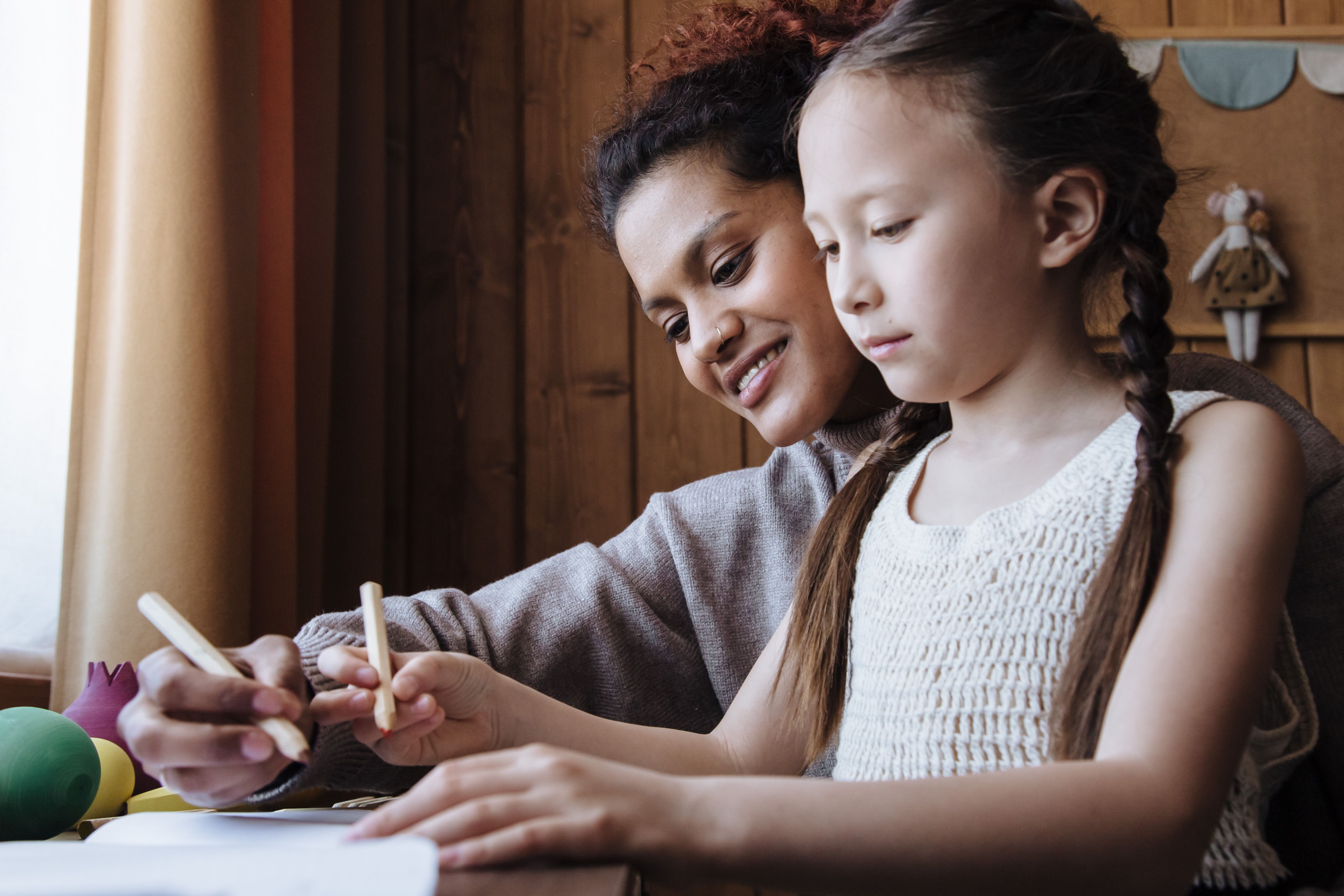 A mother and her daughter draw together at a wooden table beside a sunlit window.